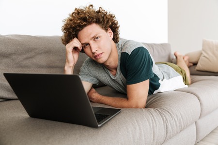 Image of handsome concentrated man lies on sofa using laptop computer. Looking camera.の写真素材