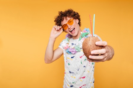 Happy young guy in sunglasses showing coconut cocktail isolated over yellow background
