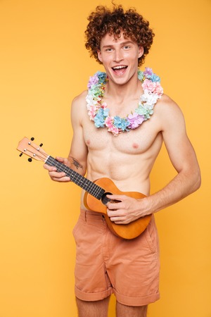 Young shirtless guy holding ukulele and looking at camera isolated over yellow backgroundの写真素材