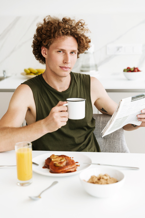 Picture of man sitting at the kitchen while holding newspaper and drinking coffee. Looking camera.の写真素材