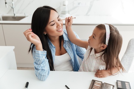 Little cute asian girl applying make up on her mothers face while sitting at the table at homeの写真素材