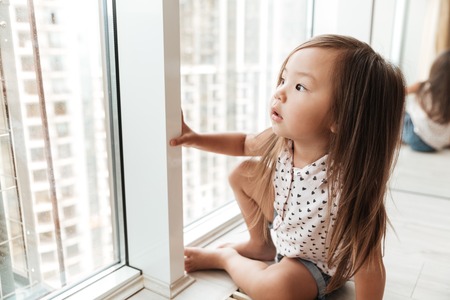 Picture of pretty little cute asian girl at home indoors standing near window. Looking aside.の写真素材