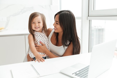 Photo of smiling young mom sitting at the table with little cute asian girl at home indoors using laptop computer writing notes to notebook.の写真素材