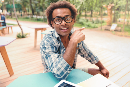 Cheerful african young man in glasses siting in cafe and pointing awayの写真素材