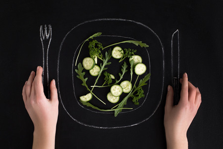 Hands holding drawn knife and fork ready to eat salad from a plate over black backgroundの写真素材
