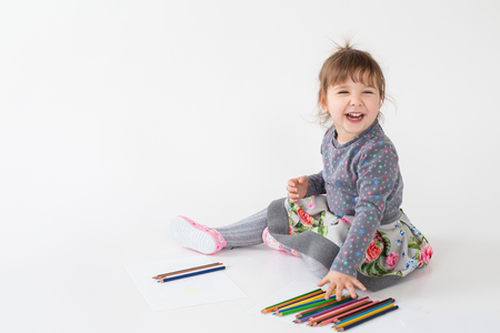 Image of happy little girl sitting near a lot of colouring pencils over white background. Looking aside.の写真素材