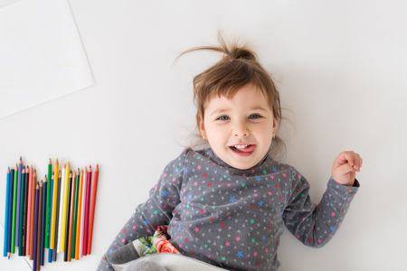 Image of cheerful little girl lies near a lot of colouring pencils over white background. Looking at camera.の写真素材