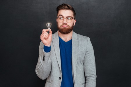 Funny amazed young businessman in glasses holding small light bulb over black backgorund