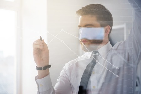 Serious bearded business man drawing business strategy on virtual screen while standing in his officeの写真素材