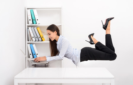Side view portrait of a smiling business woman flying above office desk and working on laptopの写真素材