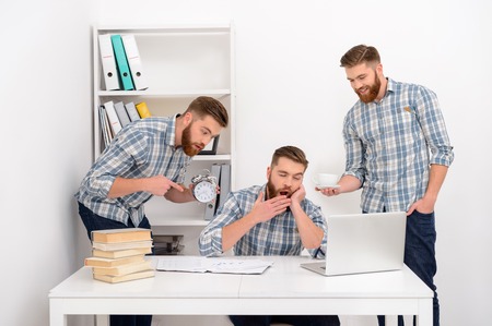 Tired sleepy businessman yawning while sitting at the table in office with two clones of himself holding alarm clock and cup of coffeeの写真素材