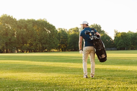 Back view of young man in cap carrying golf bag while standing on fieldの写真素材