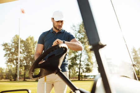 Young male golfer looking at a wristwatch while standing at the golf cartの写真素材