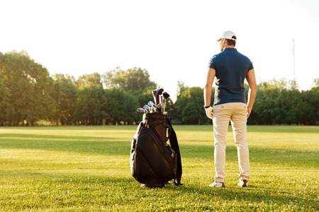 Back view of a young man standing on a green field with golf bagの写真素材