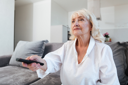 Mature woman holding tv remote control while sitting on a sofa at homeの写真素材