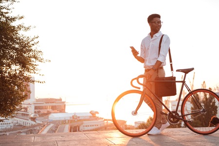 Image of young african man early morning standing near bicycle looking aside chatting by phone outdoors.の写真素材