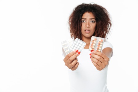 Picture of sad young african lady standing isolated over white background. Looking at camera holding pills.の写真素材
