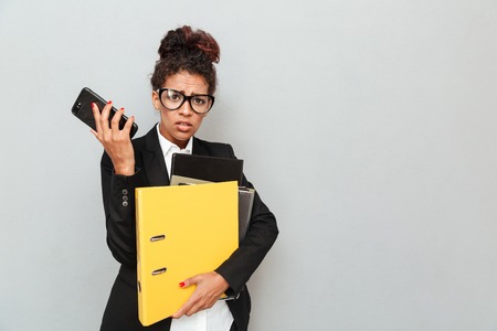 Image of confused young african business woman standing over grey wall looking camera talking by phone holding folders.の写真素材