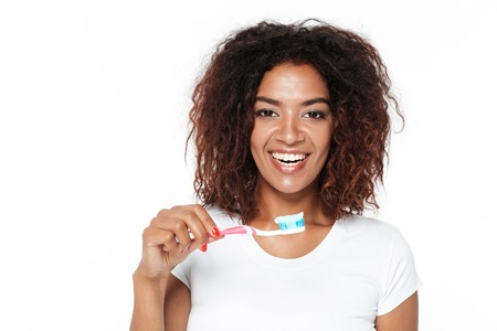 Picture of happy young african lady standing isolated over white background. Looking at camera holding toothbrush with toothpaste.の写真素材