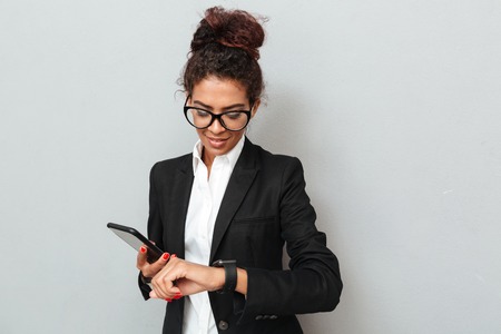 Photo of young african amazing business woman over grey wall looking aside using watch chatting by...