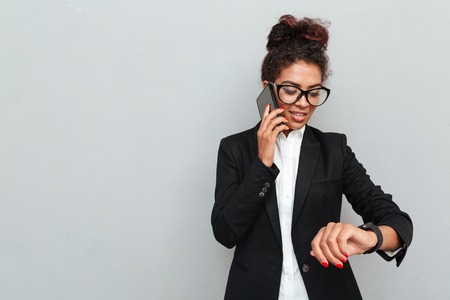 Photo of young african amazing business woman over grey wall looking aside using watch talking by phone.の写真素材