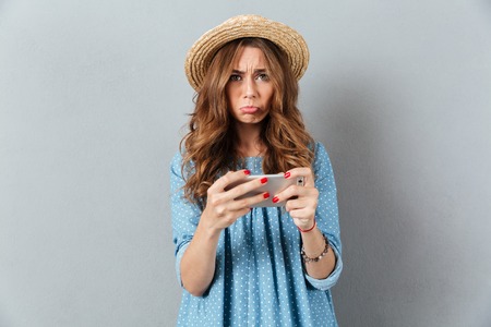 Photo of sad young caucasian woman play games by phone. Looking camera.
