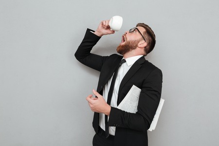 Funny young man in suit trying to drink last coffee drop from a cup isolated over gray backgroundの写真素材