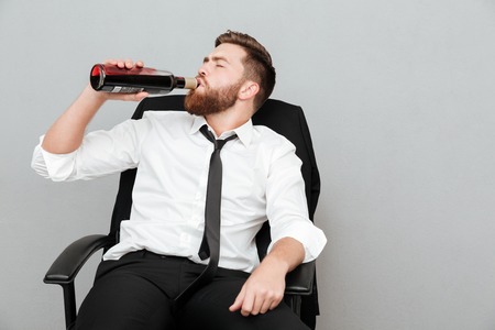 Tired young businessman sitting in office chair and drinking wine from a bottle isolated over gray backgroundの写真素材