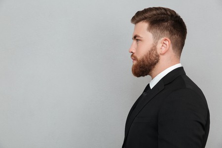 Side view portrait of a young bearded man in suit looking forward isolated over gray backgroundの写真素材