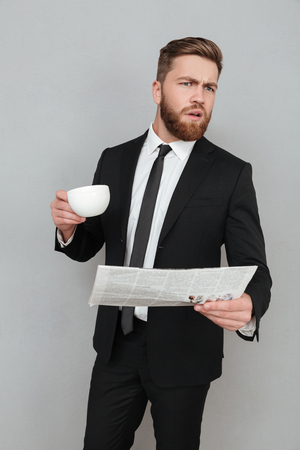 Young bearded businessman in suit holding newspaper and cup of coffee while standing isolated over gray backgroundの写真素材