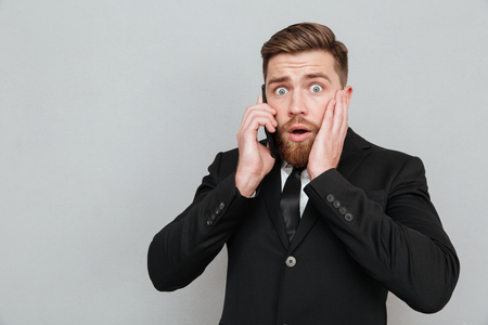 Shocked bearded man talking on his smrtphone with open mouth whil holding his cheek and looking at the cameraの写真素材