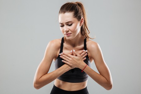 Young sportswoman having a chest pain while standing isolated over gray backgroundの写真素材