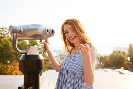 Young smiling girl standing at the telescope and gesturing with hand outdoors with a city viewの写真素材