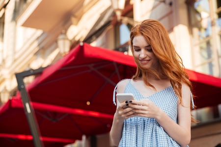 Attractive young redhead girl using mobile phone while standing outdoors on a city streetの写真素材