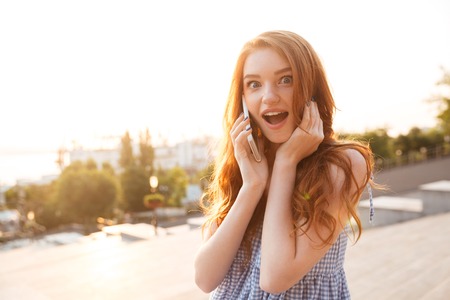 Amazed pretty redhead girl with long hair talking on mobile phone while standing over city street backgroundの写真素材