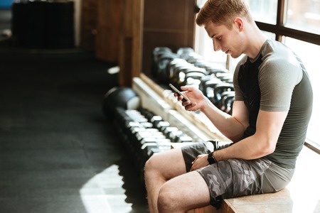 Image of caucasian sports strong man sitting in a gym chatting by phone.