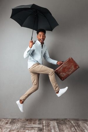 Full length portrait of a happy cheerful african man jumping with open umbrella and suitcase isolated over gray backgroundの写真素材