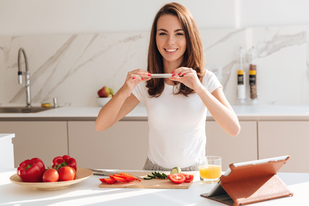 Smiling cheerful woman taking a picture of sliced vegetables on a wooden board with mobile phone while standing at the kitchenの写真素材