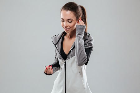 Smiling female runner in warm clothes listening music and looking down over gray backgroundの写真素材