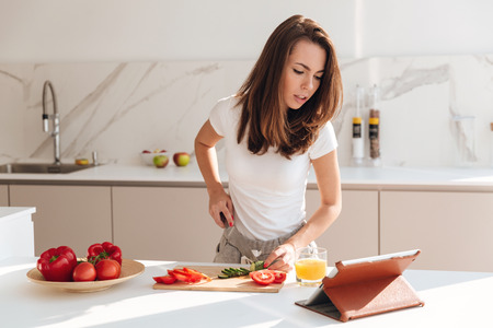 Concentrated young woman cutting vegetables on a wooden board and looking at tablet computer while cooking in a kitchenの写真素材