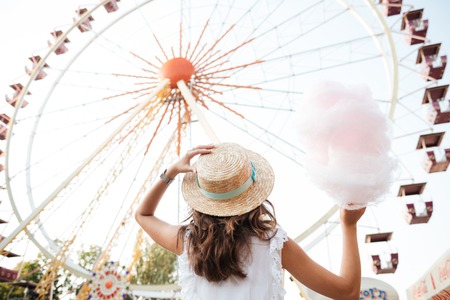 Back view of a young girl in hat holding cotton candy while standing at the ferris wheel at the amusement parkの写真素材