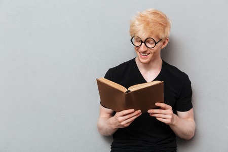 Photo of handsome cheerful caucasian man over grey wall near copyspace. Looking aside reading book.