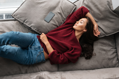 Top view of a smiling asian woman in sweater sleeping on a couch at home with blank screen mobile phone Top view of a smiling asian woman in sweater sleeping on a couch at home with blank screen mobile phone
