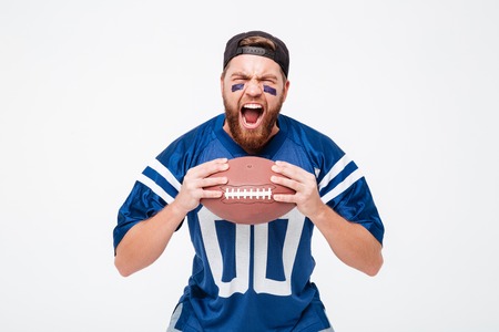Image of excited screaming man fan in blue t-shirt standing isolated over white background. Looking camera holding rugby ball.の写真素材