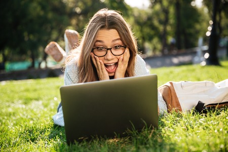 Shocked brunette woman in eyeglasses lying on grass in park and using laptop computerの写真素材