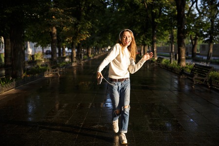 Full length portrait of happy brunette woman in autumn clothes dancing in park while listening music...
