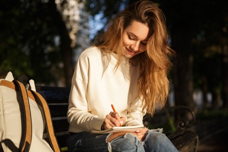 Smiling brunette woman in autumn clothes sitting on bench and writing somethingの写真素材