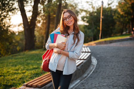 Carefree brunette woman in eyeglasses hugging books and looking at the camera in parkの写真素材