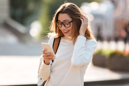 Surprised happy brunette woman in eyeglasses and autumn clothes writing message on smartphone outdoorsの写真素材