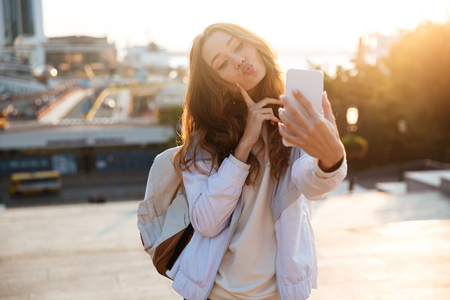 Pretty brunette woman in autumn clothes making selfie on smartphone and showing grimace outdoorsの写真素材